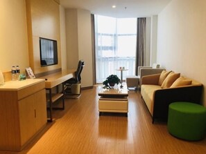 Une chambre d’hôtel moderne avec un plancher en bois, un canapé, une table basse, une télévision, un bureau avec une chaise et une fenêtre avec des rideaux.
