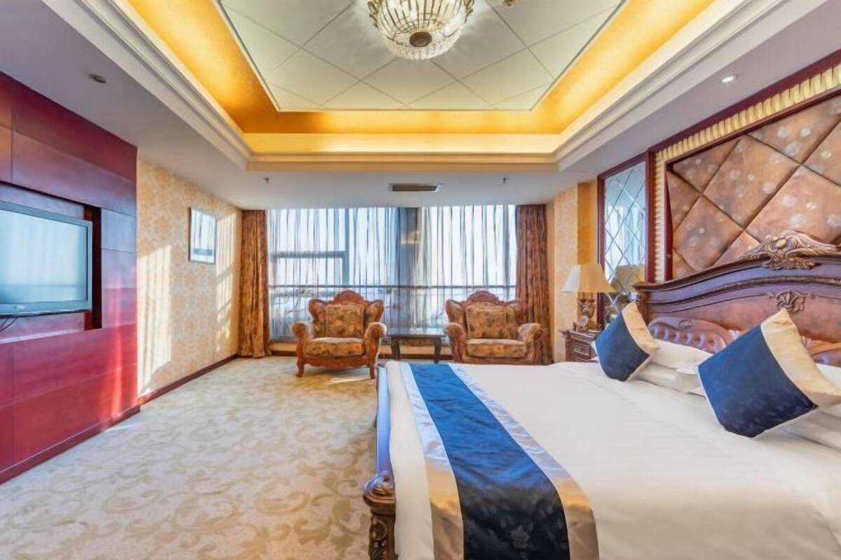 A hotel room with a large bed, two armchairs, a TV, and a chandelier.
