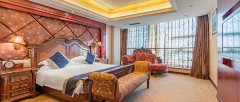 A hotel room with a large bed, a wooden nightstand, a bench, and a view of the outdoors through large windows.