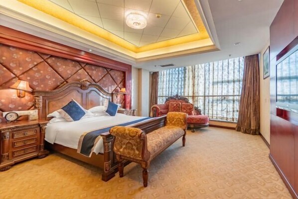 Quarto de hotel com uma cama grande, uma mesa de cabeceira de madeira, um banco e vista para o exterior através de amplas janelas.