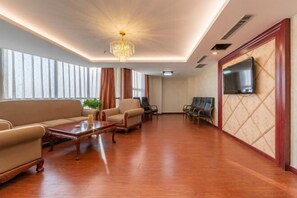 Uma sala de estar ampla com piso de madeira, um sofá, poltronas, uma mesa de centro, um lustre e uma TV fixada na parede.