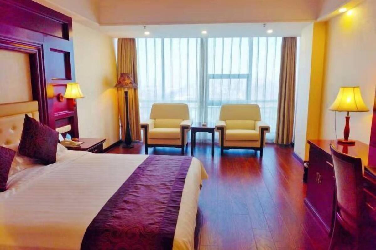 A hotel room with a large bed, two armchairs, a desk, and a view of the city through large windows.