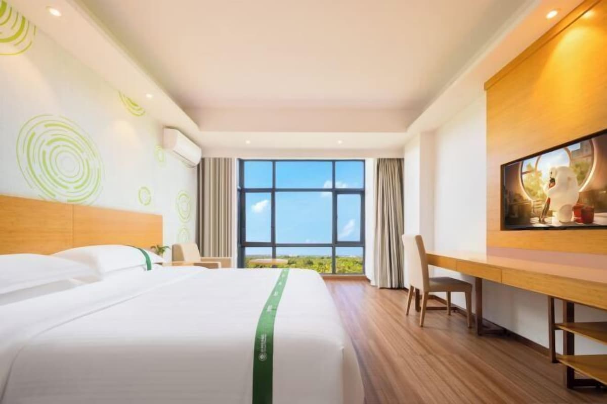 A hotel room with a large bed, a desk, a chair, and a large window offering a view of the outdoors.