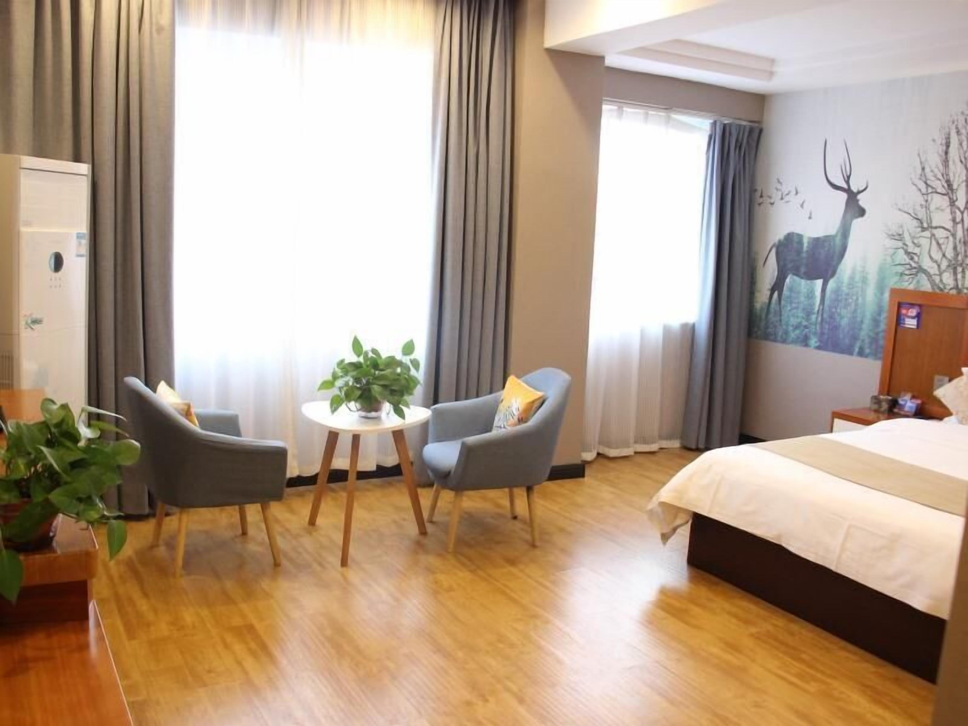 A hotel room with a bed, two armchairs, a small table with a plant, and a large window with curtains.