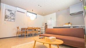 Espaço de estar compacto com sofá, área de refeições com cadeiras, cozinha equipada e iluminação embutida no teto.