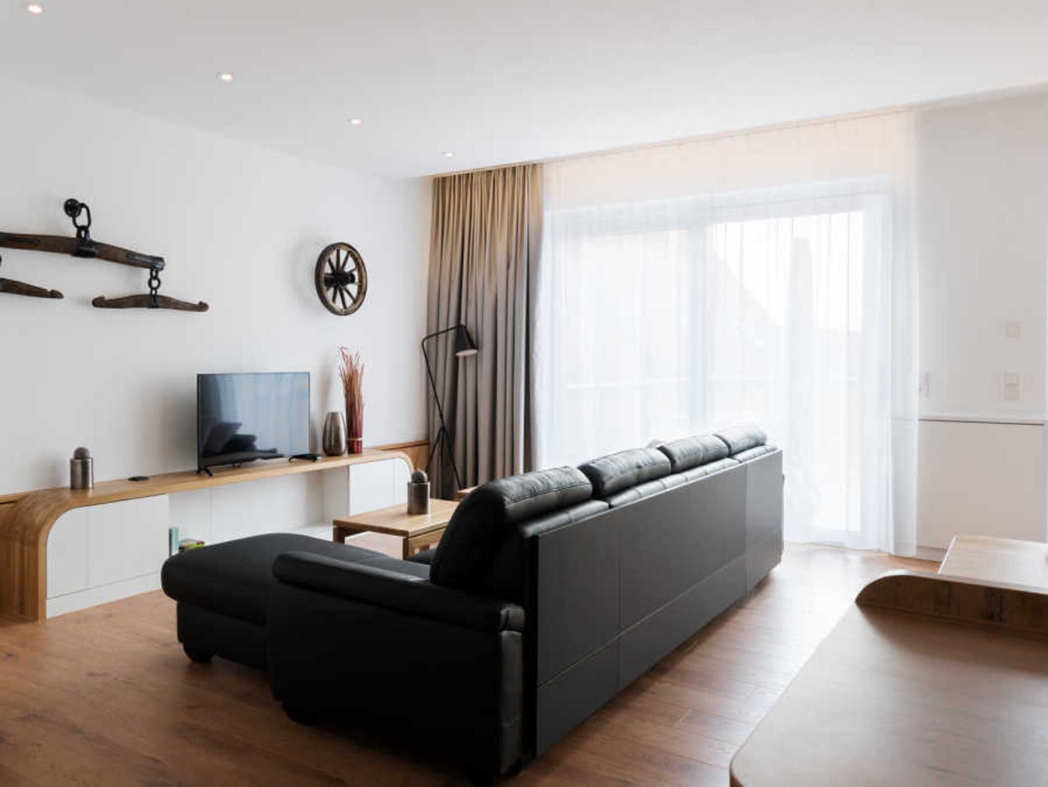 En moderne stue med en sort sofa, et træbord, et fjernsyn og et stort vindue med gardiner.