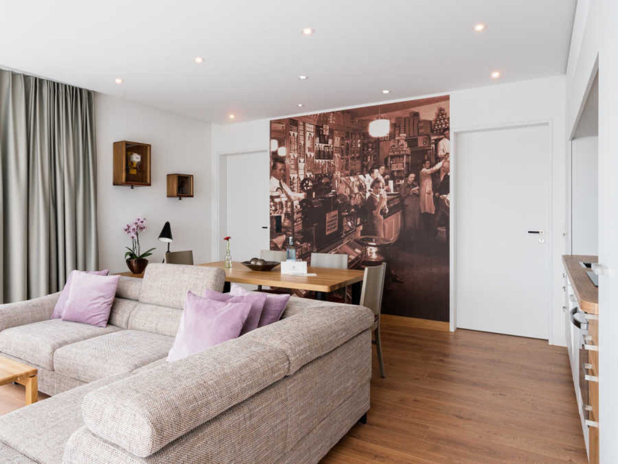 Een moderne woonkamer met een bank, een eettafel en een groot muurschildering dat een levendige barscene voorstelt.