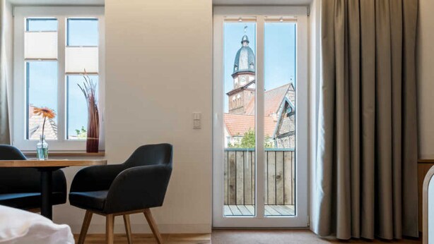 Hotelkamer met een houten vloer, een bed, een bureau met stoel, een raam met uitzicht op een gebouw en een schuifdeur naar een buitenbalkon.