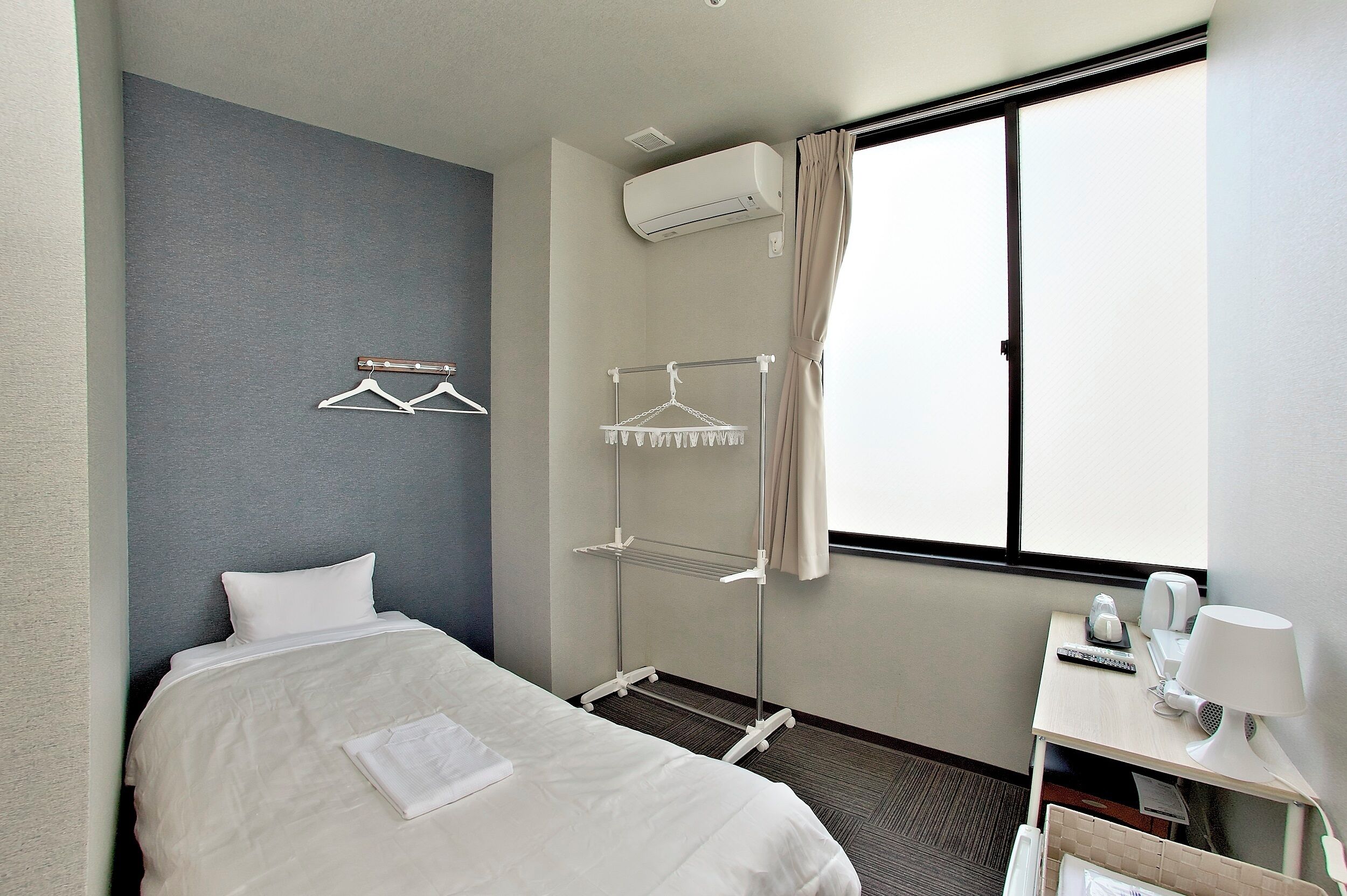 A hotel room with a bed, a bedside table with a lamp, a desk with a chair, a window with curtains, and a wall-mounted air conditioner.