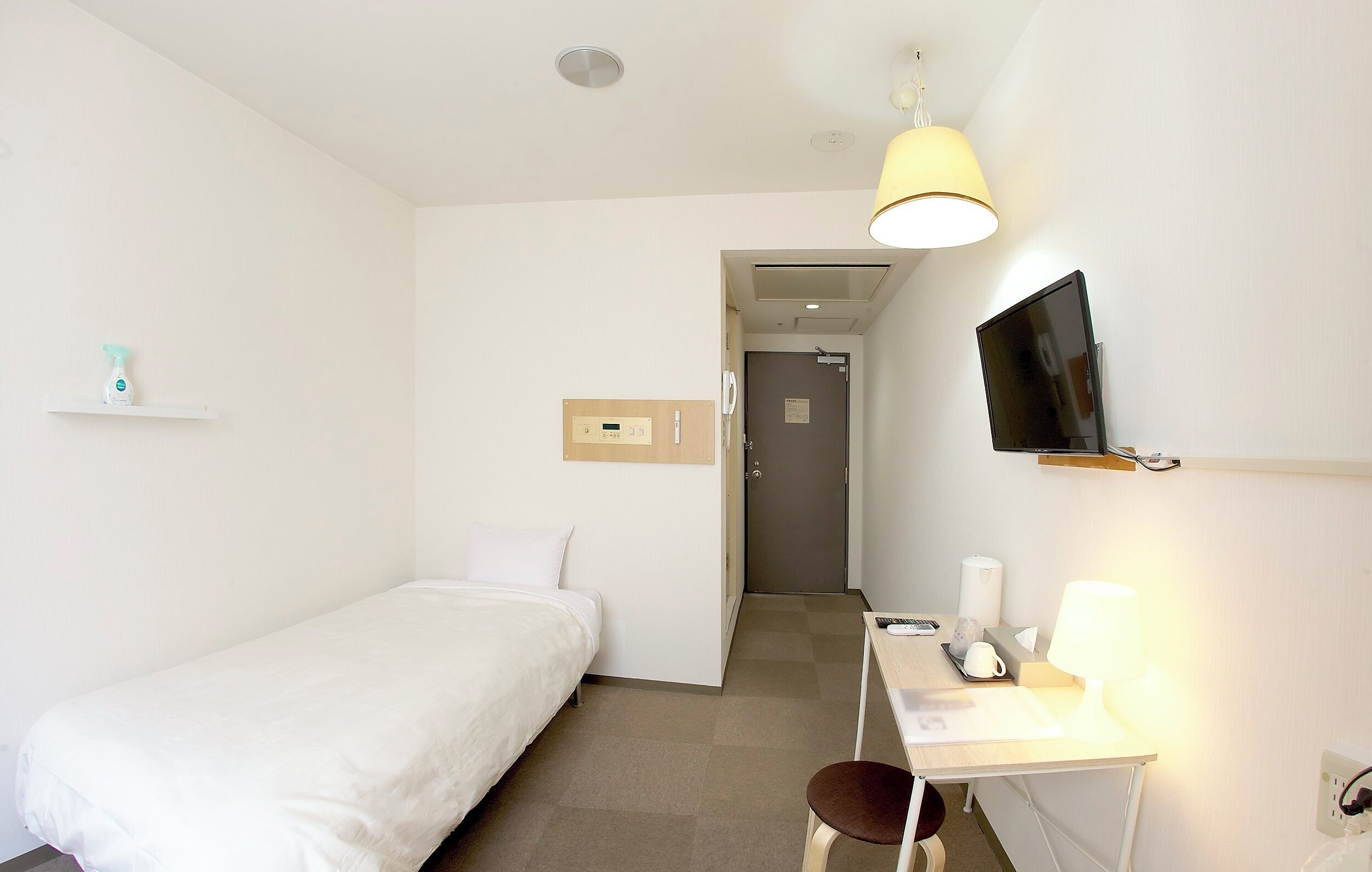 A hotel room with a single bed, a desk with a lamp, a TV mounted on the wall, and a door leading to a bathroom.