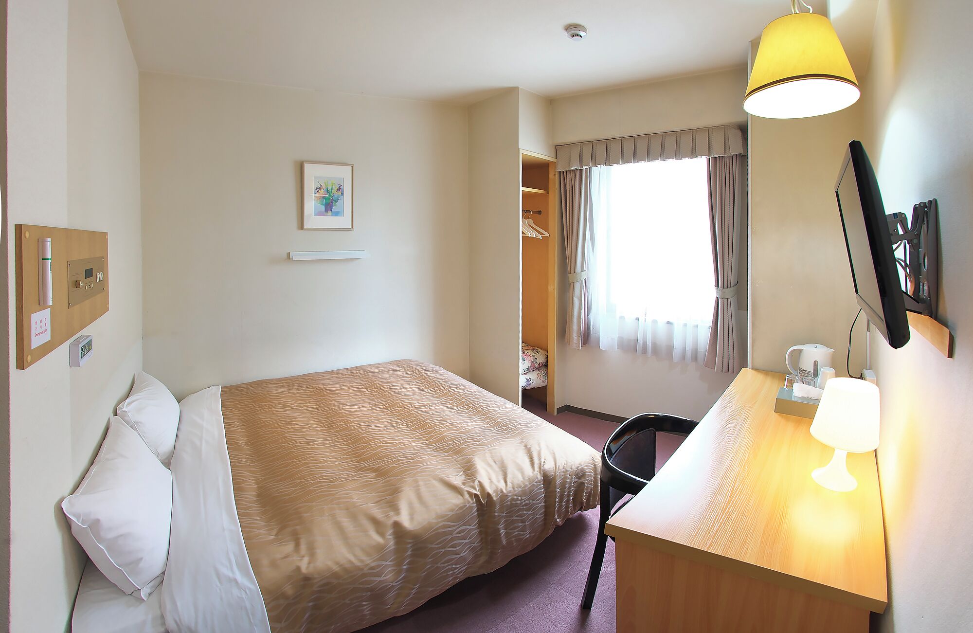 A hotel room with a bed, a desk with a lamp, a chair, a wardrobe, and a window with curtains.