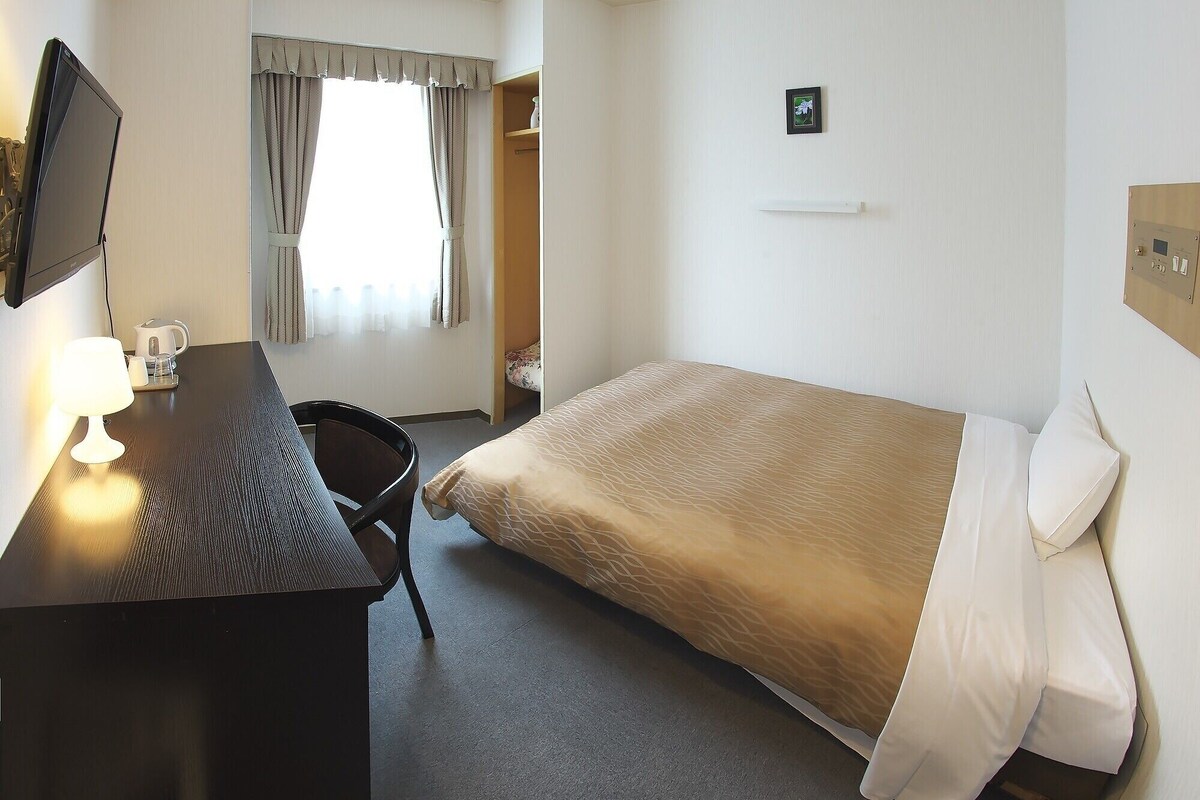 A hotel room with a bed, a desk with a computer monitor, a chair, and a window with curtains.
