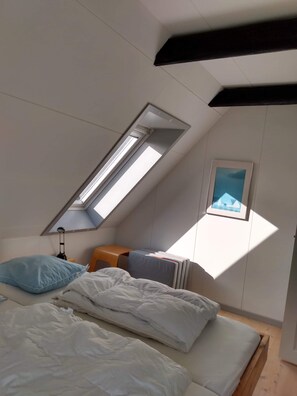 Une chambre avec un plafond en pente, un puits de lumière, un lit avec une literie blanche, un bureau avec une chaise et un tableau encadré au mur.