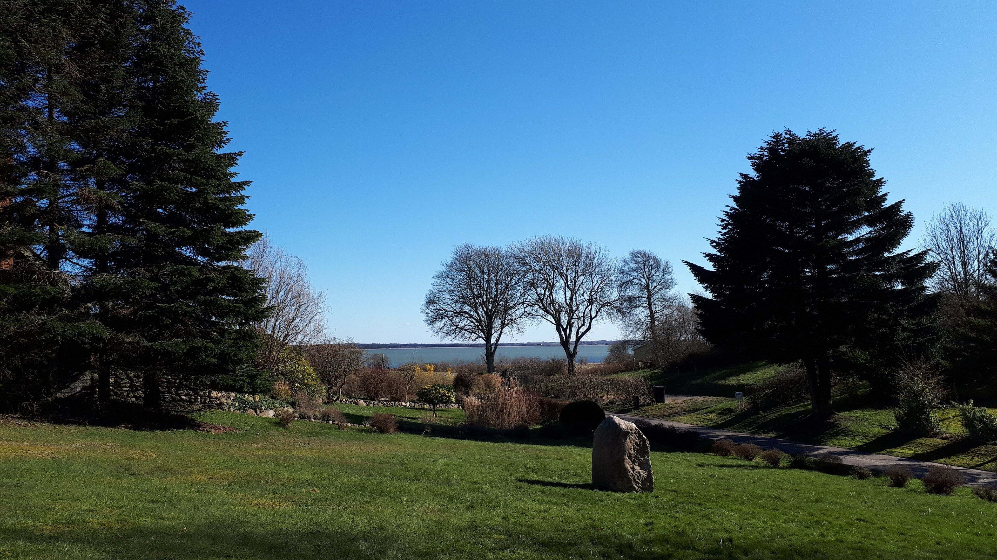 A grassy area with a large rock, trees, and a distant view of a body of water.