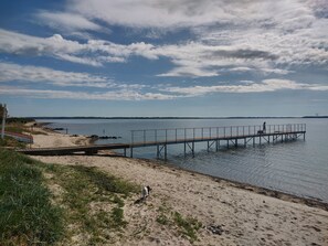 A beach with a wooden pier extending into calm waters, a sandy shore, and a clear sky with clouds. - Holiday home with views and close to water (Øster Assels)