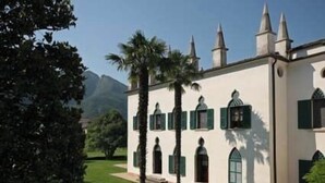 Un edificio blanco con contraventanas verdes y numerosas ventanas, rodeado de un jardín cuidado y palmeras.