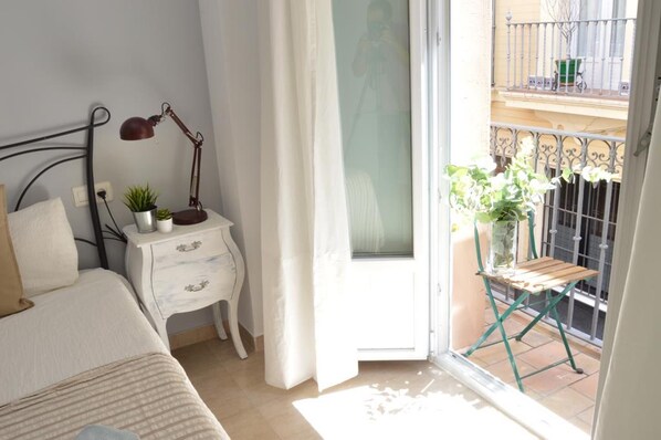 Ein Schlafzimmer mit Bett, Nachttisch mit Lampe, einer kleinen Pflanze und einem Fenster mit Blick auf einen Balkon.