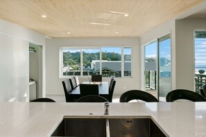 A modern dining area with a large table and chairs, large windows offering a view of a coastal landscape.