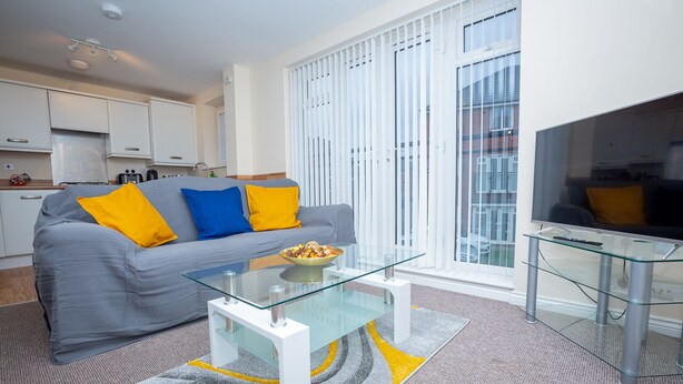 A modern living room with a grey sofa, yellow and blue cushions, a glass coffee table, and a TV on a glass stand. - Grande Richesse Apartment (Coventry)