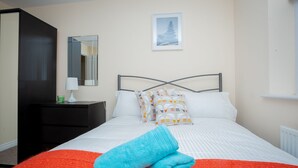 A neatly made bed with white bedding and a blue towel, a black nightstand with a lamp, and a framed picture on the wall. - Grande Richesse Apartment (Coventry)