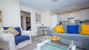 A modern living room with a glass coffee table, white sofas with colorful cushions, and a kitchen in the background. - Grande Richesse Apartment (Coventry)