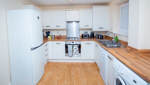 A compact kitchen with white cabinets, a stainless steel range hood, a built-in oven, a dishwasher, and a washing machine. - Grande Richesse Apartment (Coventry)