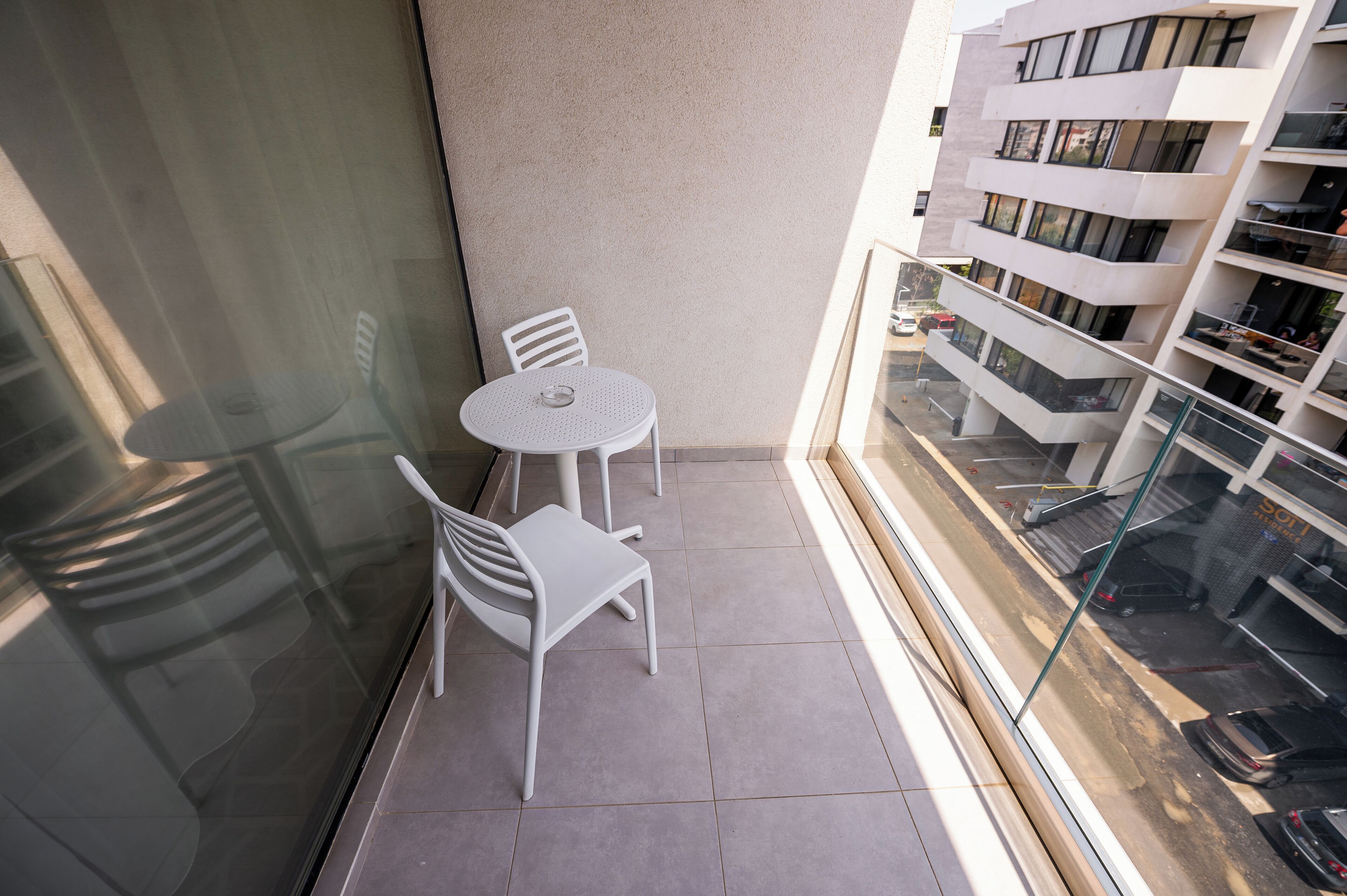 Uma varanda com uma mesa pequena e duas cadeiras, guarda-corpo de vidro e vista de um edifício com vários andares.