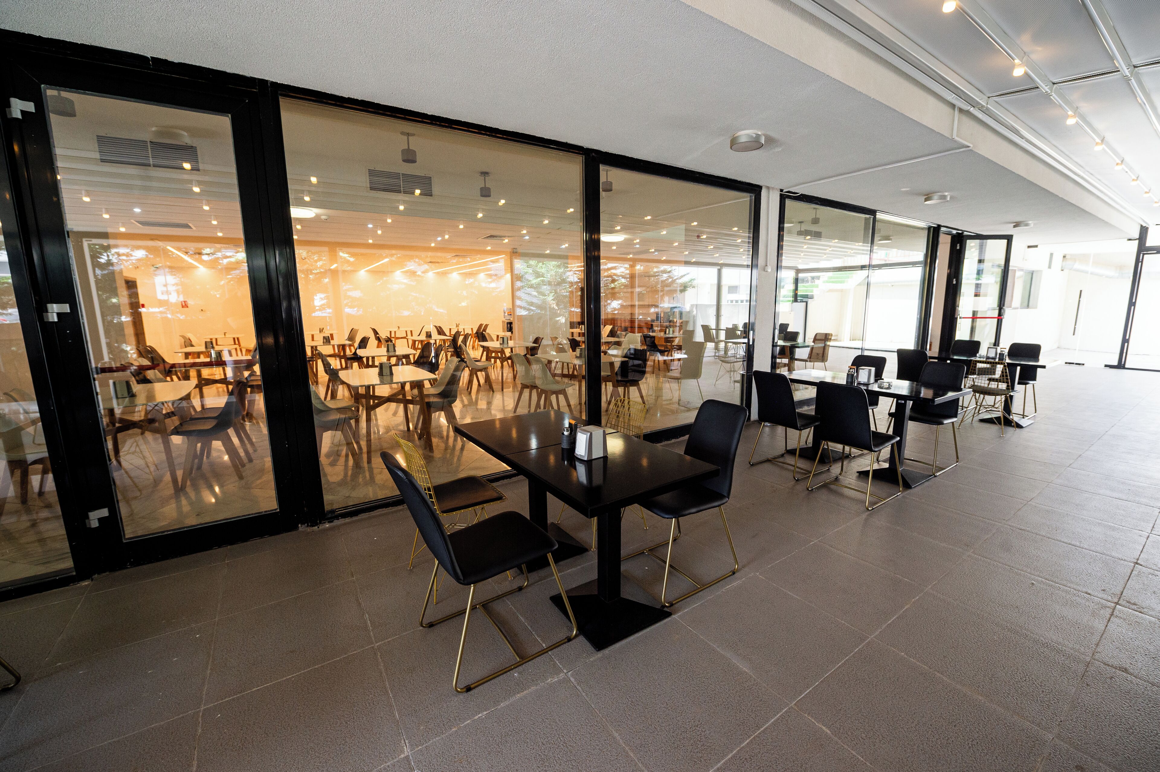 Een ruime, overdekte eetruimte met meerdere tafels en stoelen, grote ramen en een modern plafond.