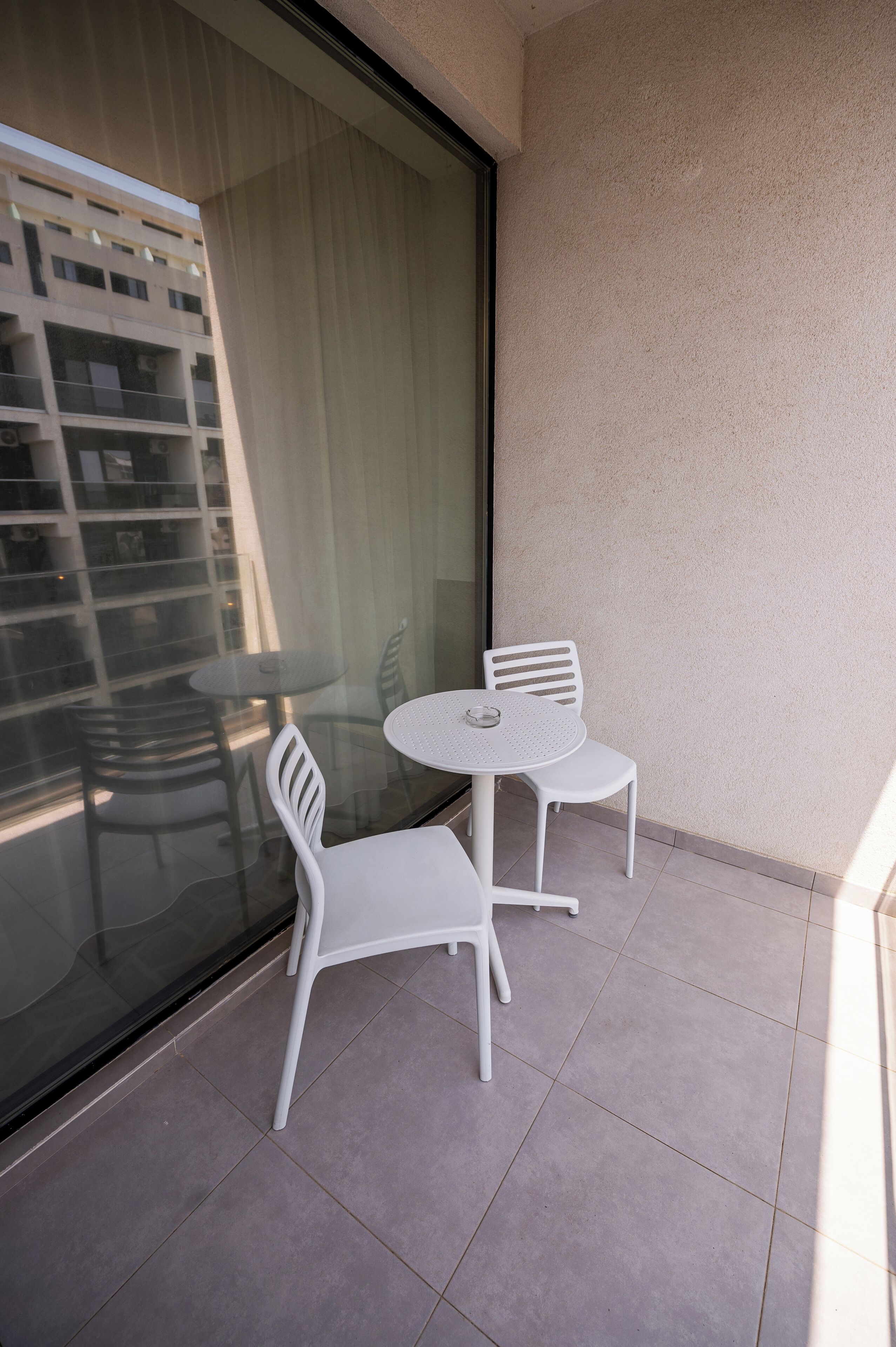 A balcony with two white chairs and a small round table, a glass door, and a view of a building with multiple floors.