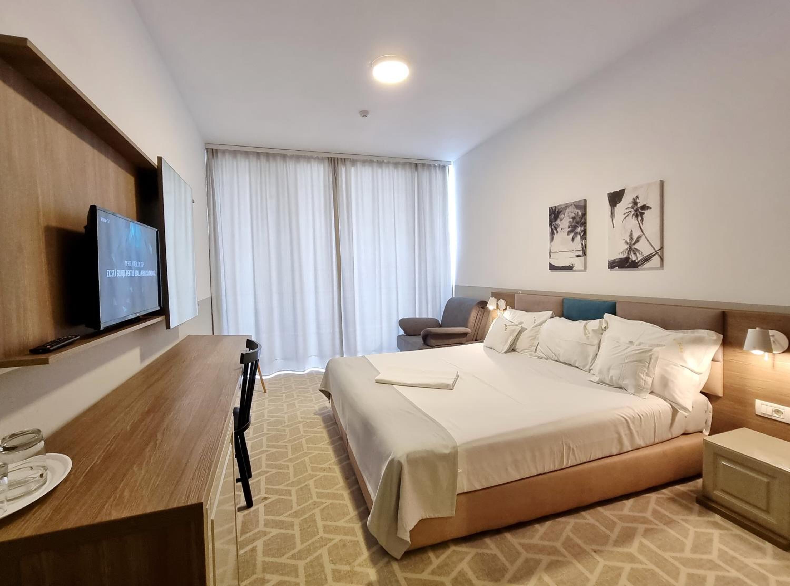 A hotel room with a bed, a desk with a TV, a chair, and a window with curtains.