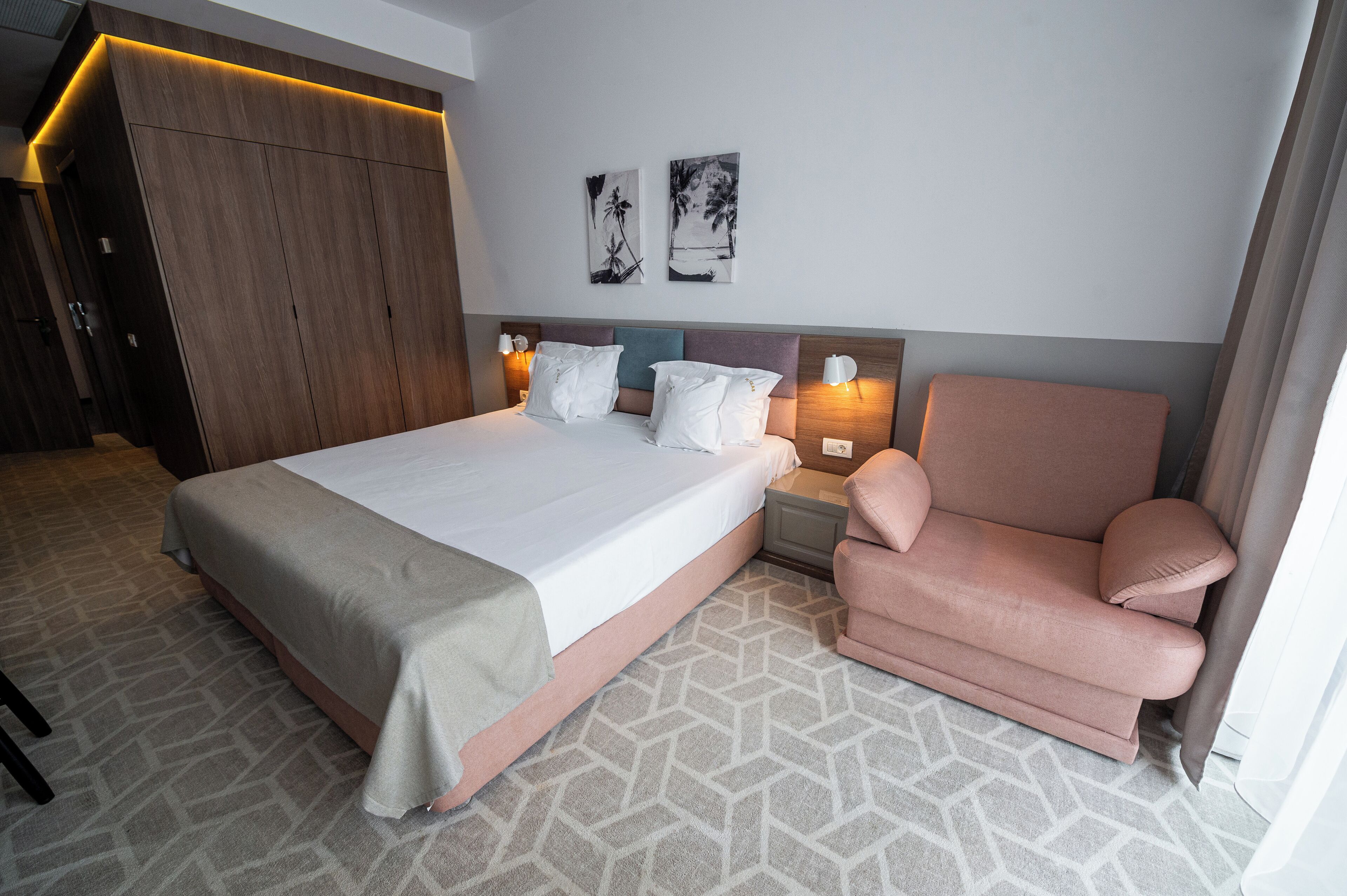 A hotel room with a large bed, a pink armchair, and two wall-mounted nightstands with lamps.
