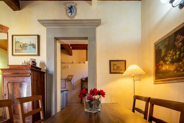 A dining area with a wooden table, chairs, a vase with flowers, a lamp, and a painting on the wall.