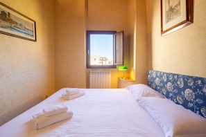 A hotel room with a bed, a window with a view, a radiator, and a green lamp on a bedside table.