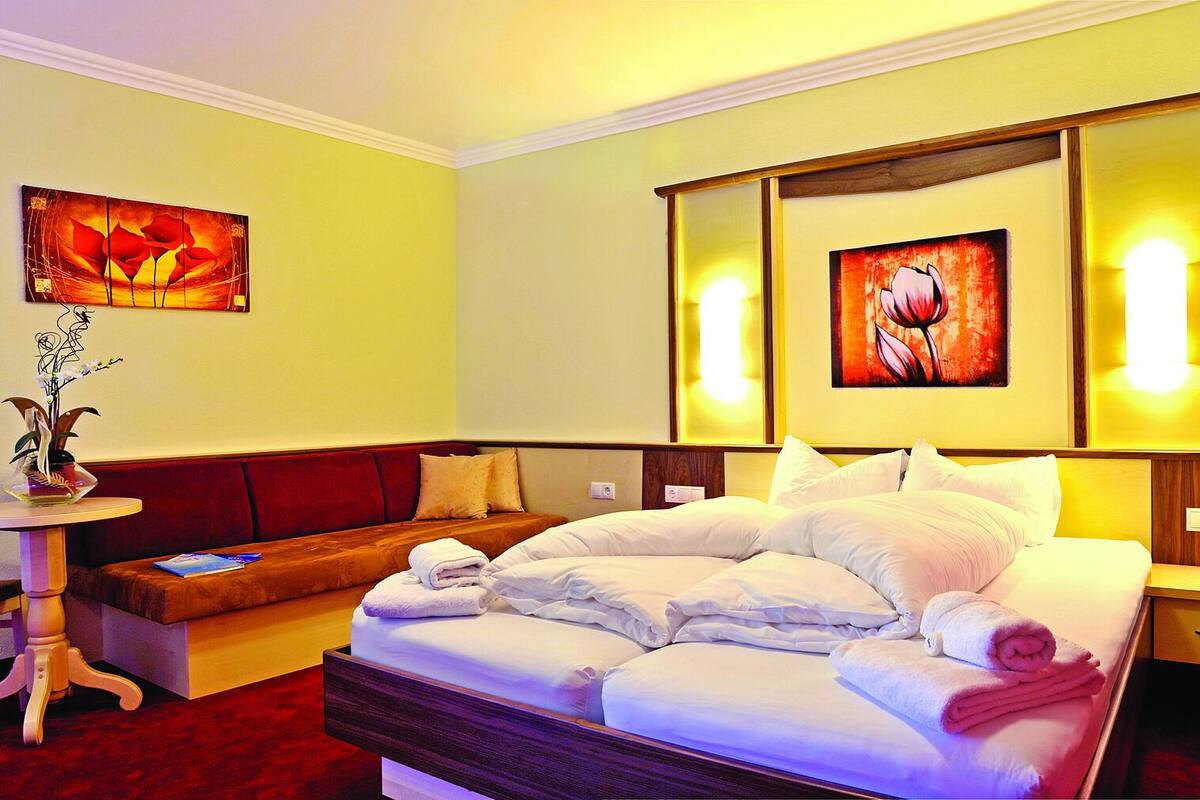 A hotel room with a bed, a sofa, a small table with a vase, and two paintings on the walls.
