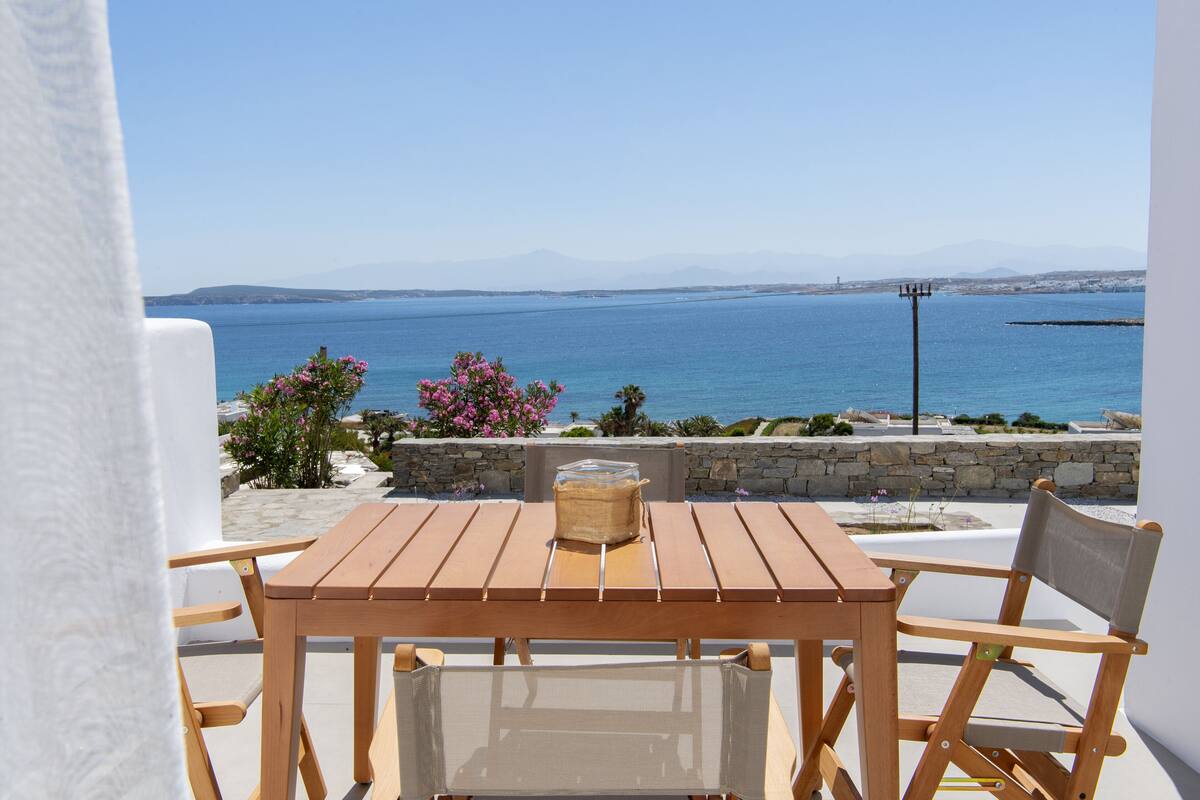 A wooden table and chairs on a balcony with a view of the sea and distant mountains.