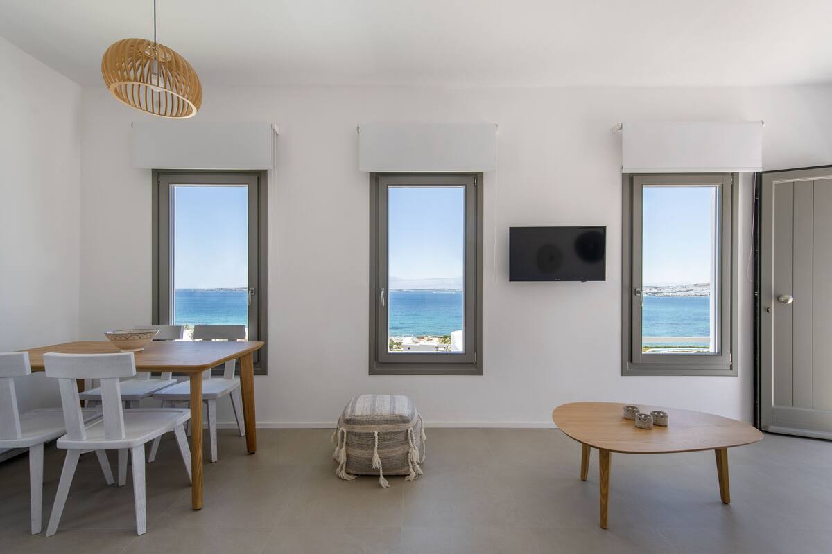 A room with a wooden table, white chairs, a small stool, and a round table with a few items on it. There are three windows providing a view of the sea.
