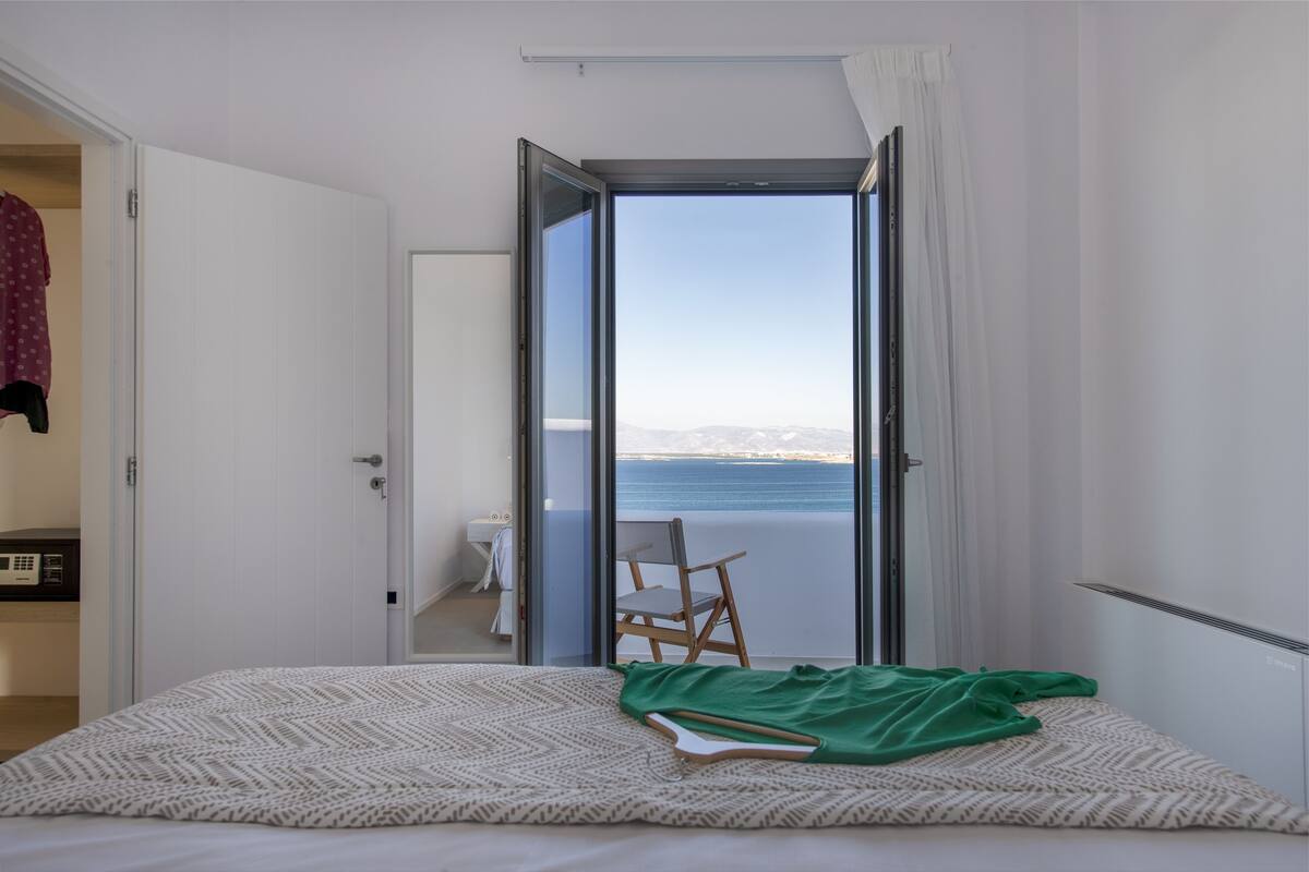 A bedroom with a bed, a chair, and a view of the sea through an open door.