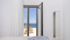 A bedroom with a bed, a large window, and a view of the beach. - Kymo Luxury Suites Paros (Paros)
