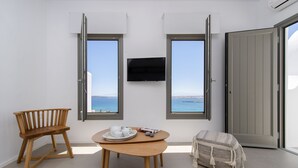 A room with a wooden bench, a small round table with a tray of white cups and saucers, and a woven pouf. Two windows with open shutters provide a view of the sea. - Kymo Luxury Suites Paros (Paros)