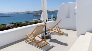 A balcony with two wooden chairs and a white umbrella, overlooking a coastal view with buildings and water. - Kymo Luxury Suites Paros (Paros)