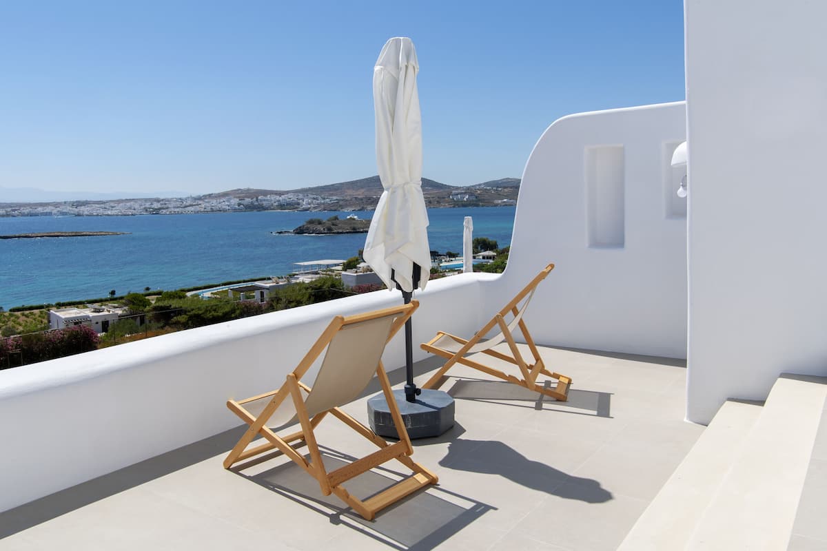 A balcony with two wooden chairs and a white umbrella, overlooking a coastal view with buildings and water.