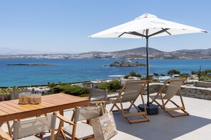 A terrace with a wooden table, two chairs, and a large umbrella overlooking a coastal view with clear blue waters and distant hills. - Kymo Luxury Suites Paros (Paros)