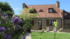 A stone house with a red-tiled roof, a stone chimney, and a small garden with purple flowers.