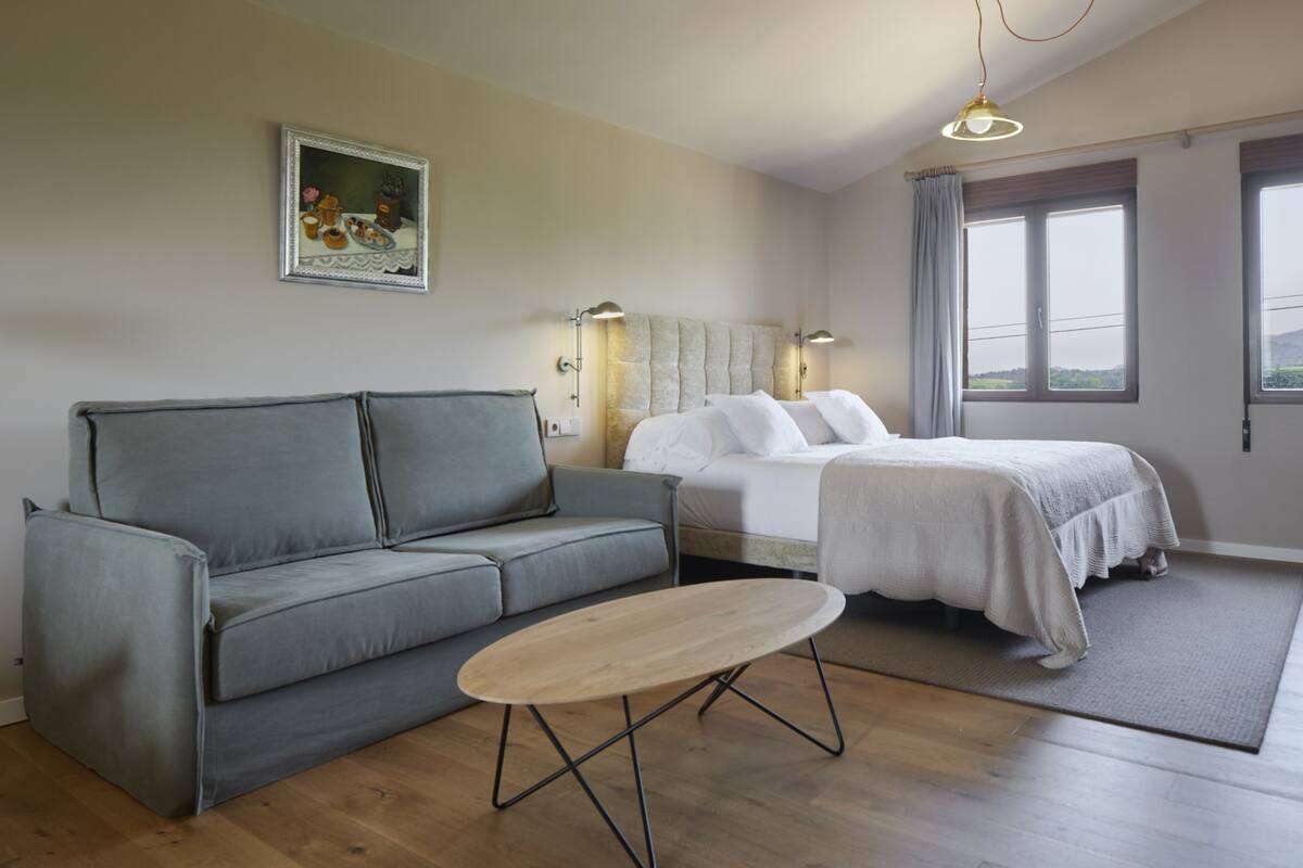 Ein modernes Schlafzimmer mit einem grauen Sofa, einem Holztisch, einem Bett mit weißer Bettwäsche und einem Gemälde an der Wand.