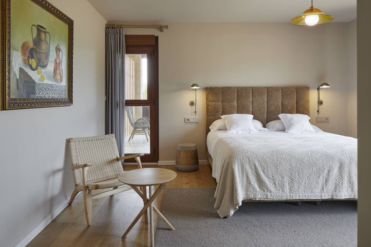 Ein Schlafzimmer mit einem großen Bett, zwei Nachttischlampen, einem Sessel, einem kleinen Tisch und einem Gemälde an der Wand.