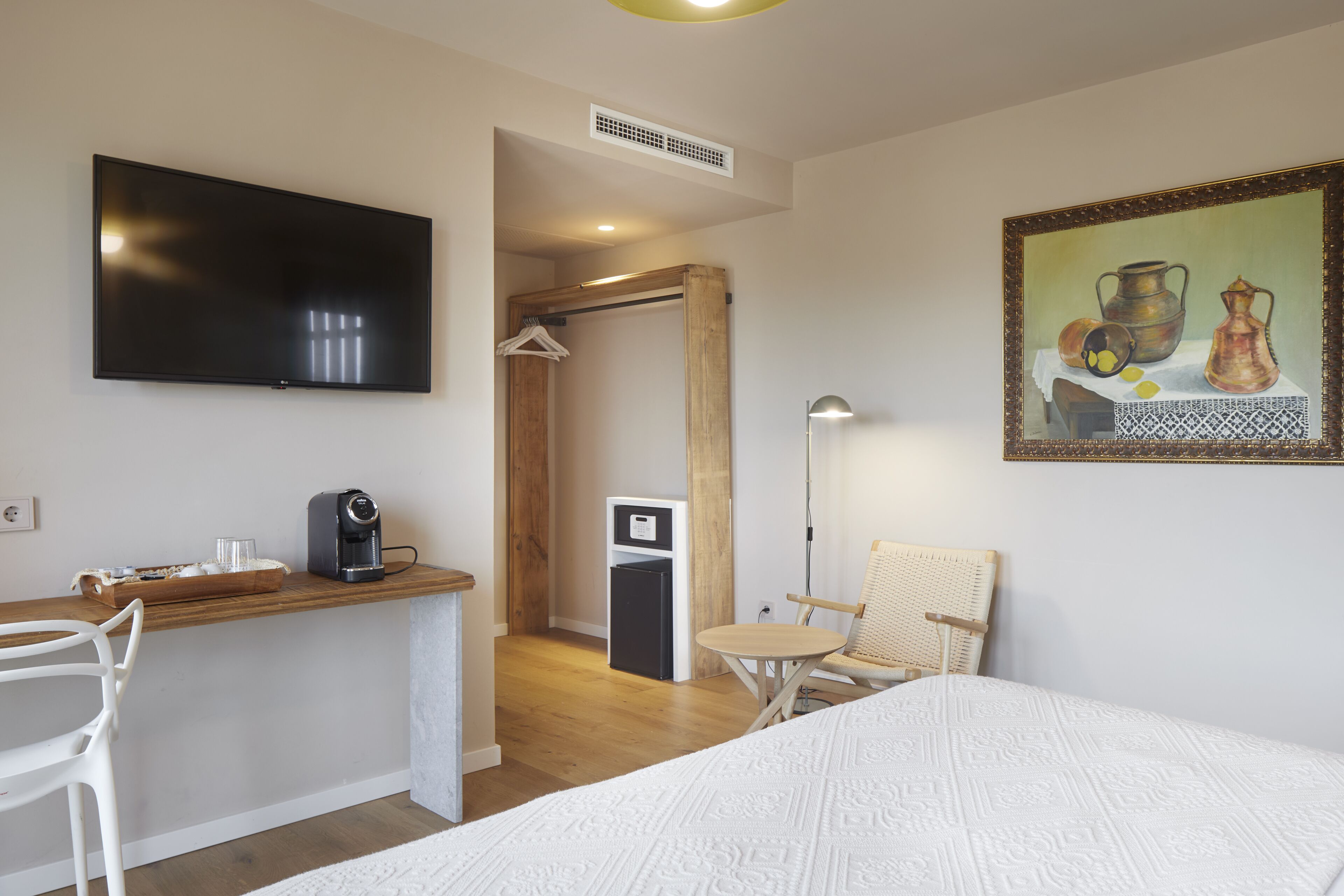 A hotel room with a bed, a desk with a coffee maker, a TV, a chair, and a painting on the wall.