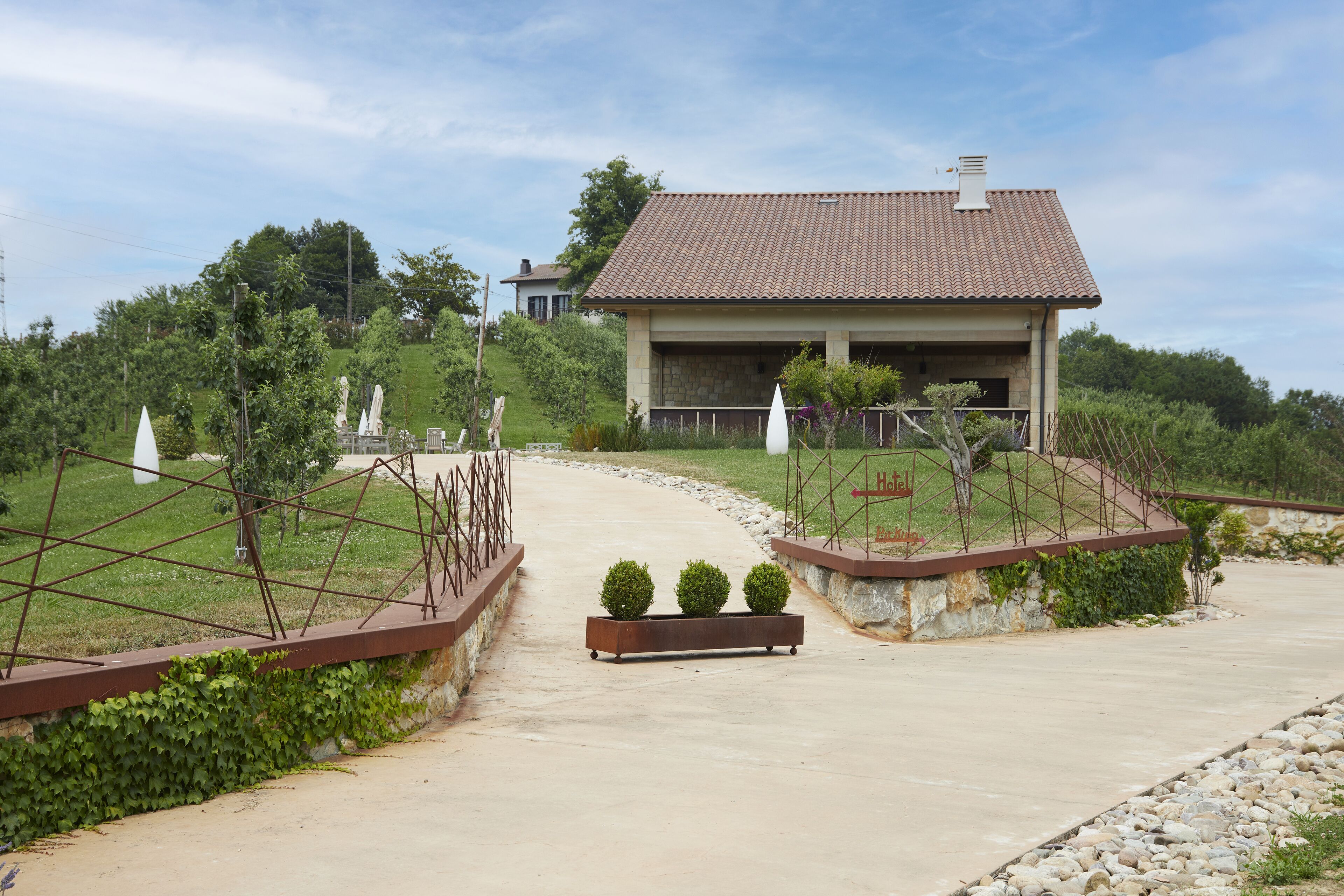 A house with a tiled roof, a paved driveway, and a garden with a bench and two spherical plants.