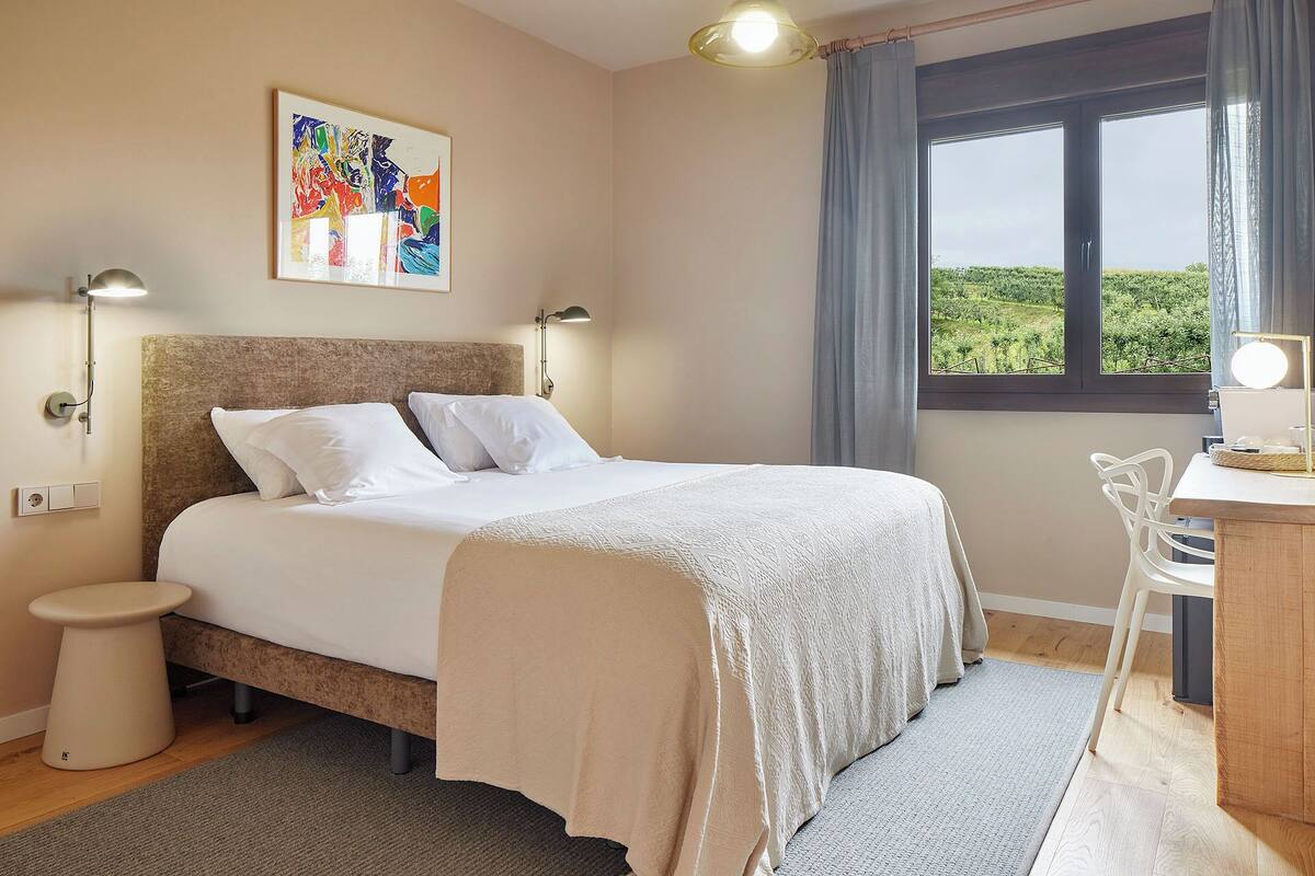 Ein ordentlich eingerichtetes Schlafzimmer mit Bett, Nachttischlampen, einem Schreibtisch und einem Fenster mit Blick ins Grüne.