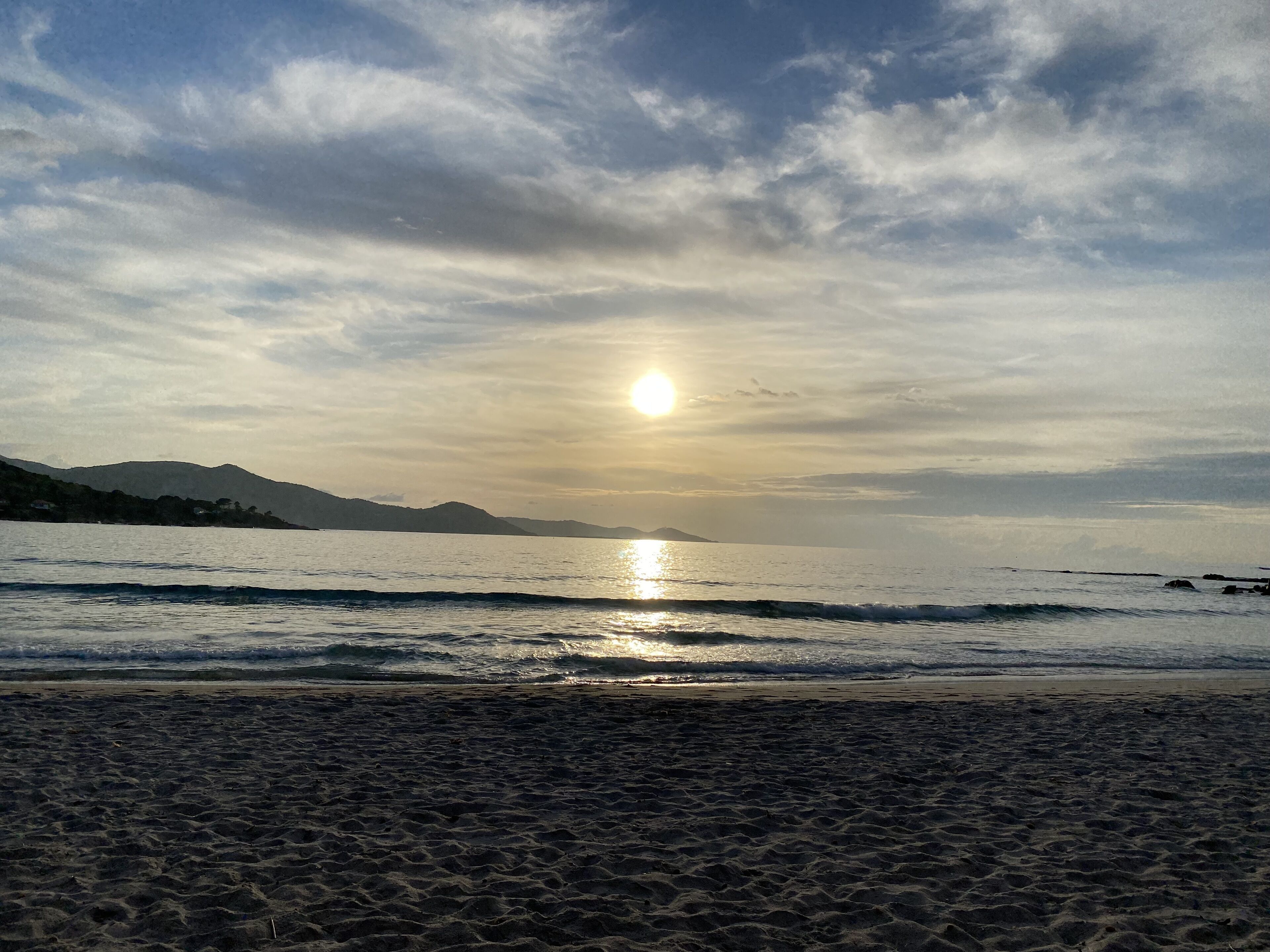 Une plage où l’on peut admirer le coucher de soleil sur l’eau, avec des montagnes en arrière-plan et une rive sablonneuse.