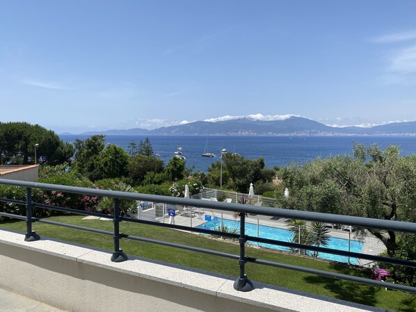 A balcony with a view of a swimming pool, a mountainous landscape, and the sea.